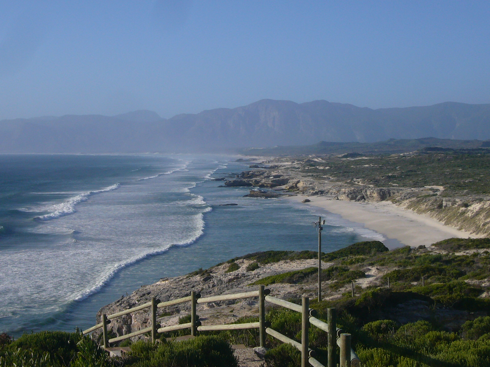 A timber boardwalk above the long white beach below De Kelders, with the mountains of Walker Bay beyond